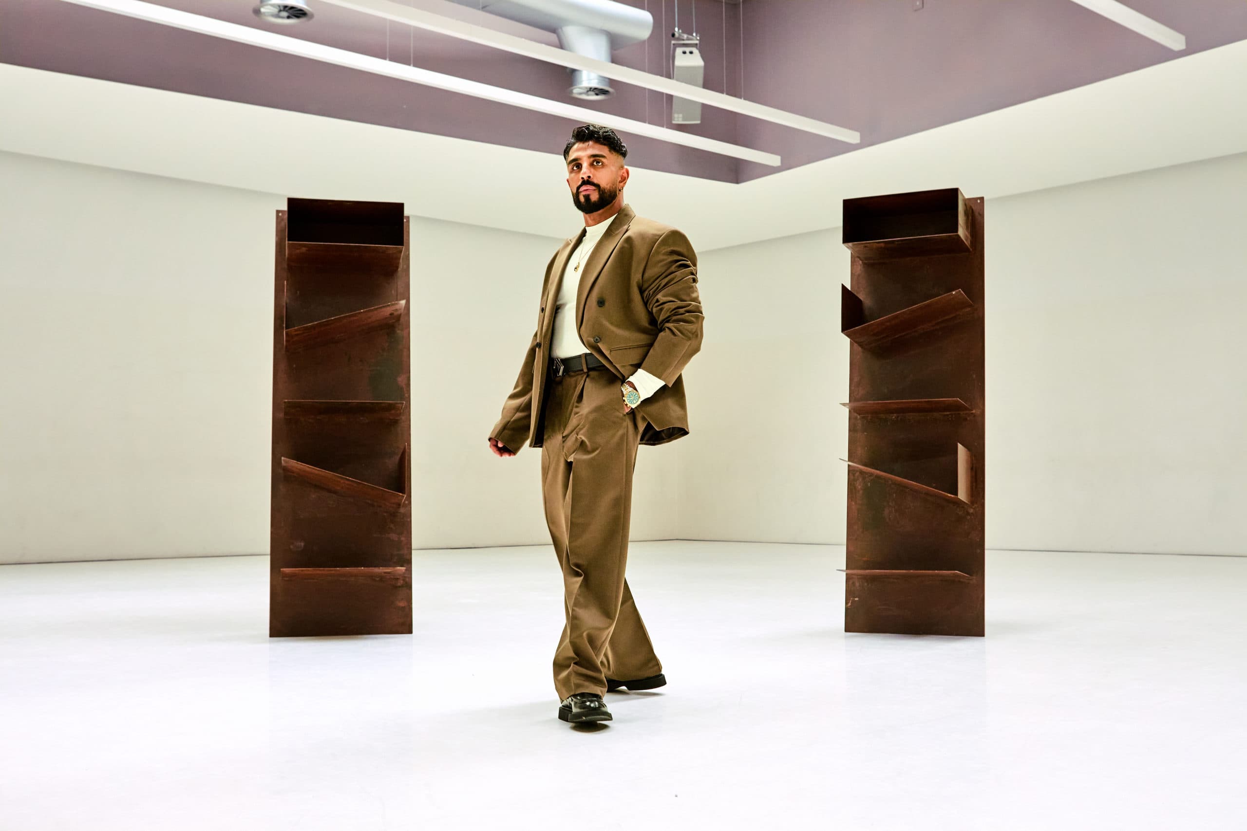 Omar Bhatti standing beside a tall rust-coloured steel sculpture in a white gallery